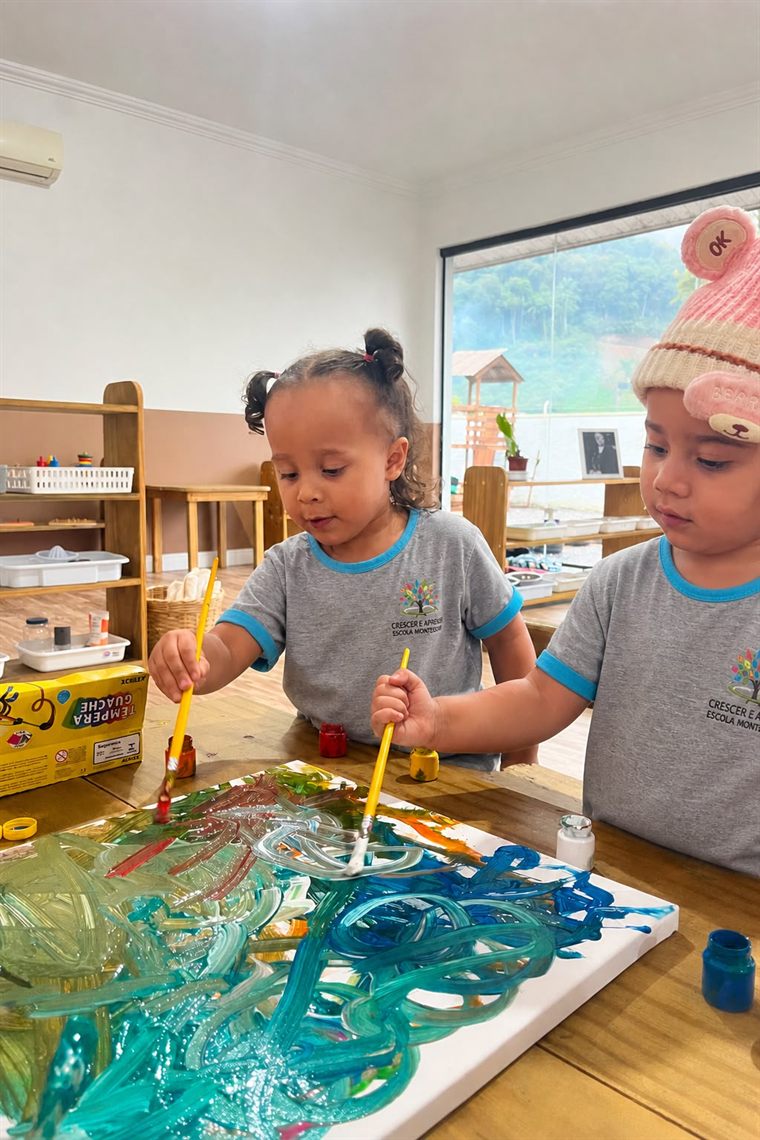 Duas crianças pintando com pincéis e tinta guache sobre uma tela, em uma sala Montessori
