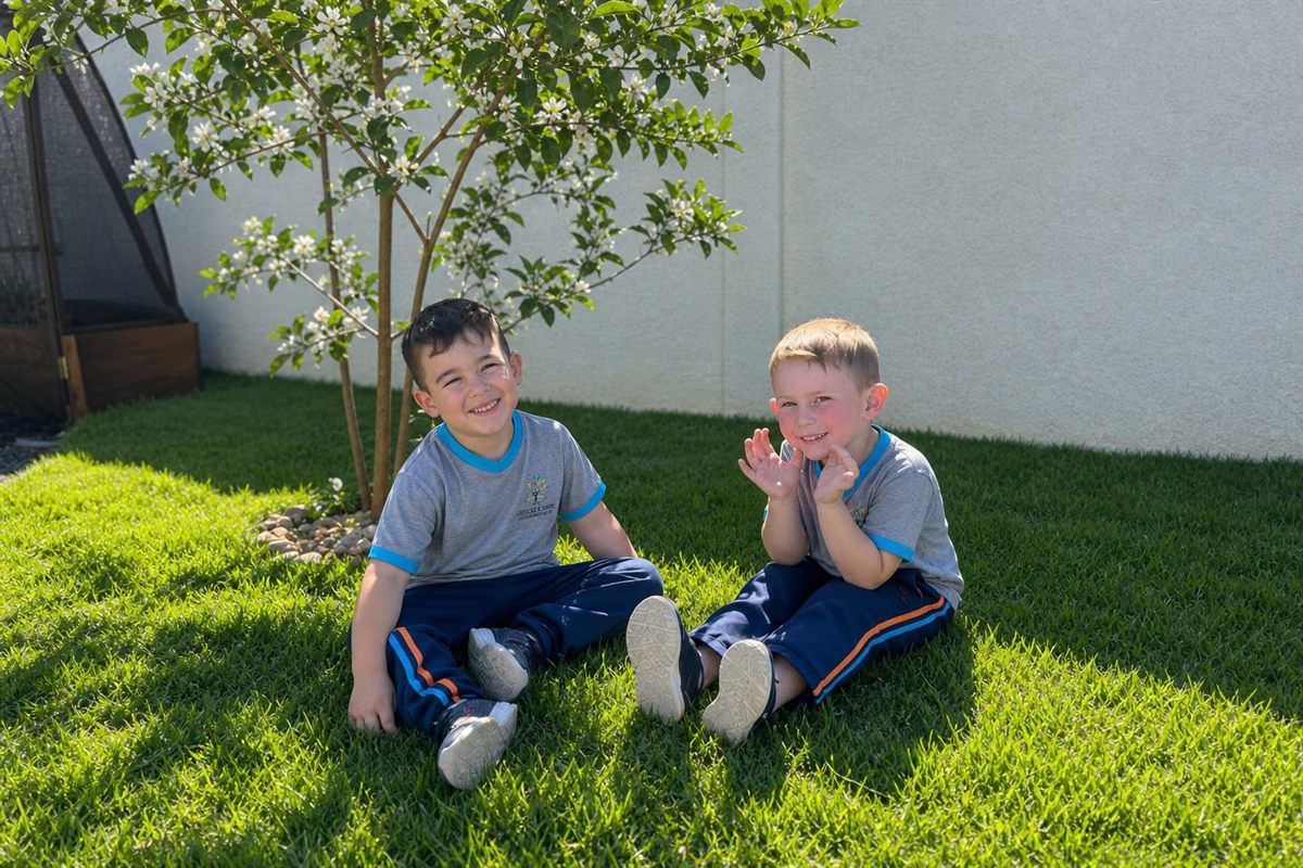 Dois meninos sorrindo, sentados na grama do jardim da escola, ao lado de uma árvore florida